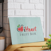The iHeartFruitBox Glass Cutting Board sits on a table, accompanied by a potted plant.