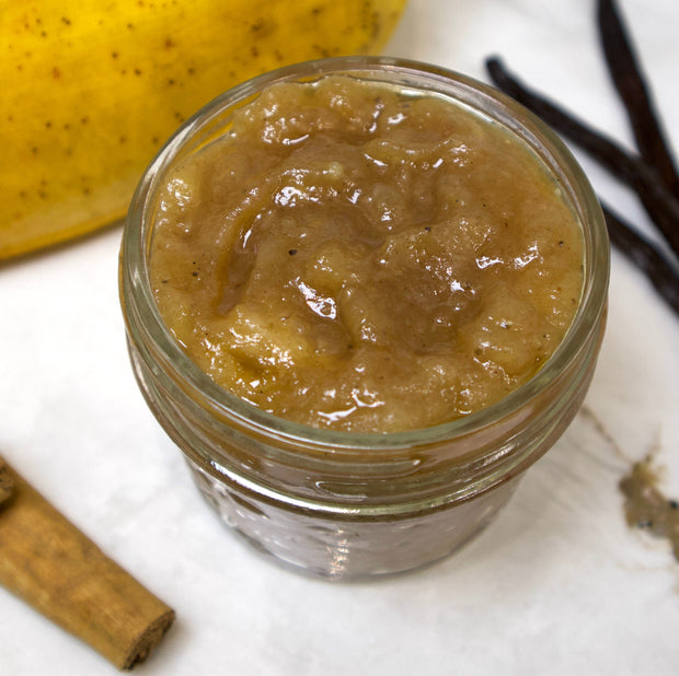 A small glass jar of iHeartFruitBox’s Going Bananas Preserve, made from rescued fruit, sits on a white surface next to a cinnamon stick, a vanilla bean, and part of a yellow apple.