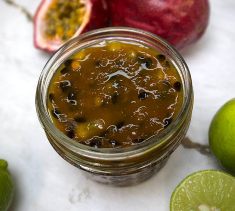 A glass jar of iHeartFruitBox Passion Fruit Preserve sits on a marble surface, surrounded by whole and halved passion fruits and limes.
