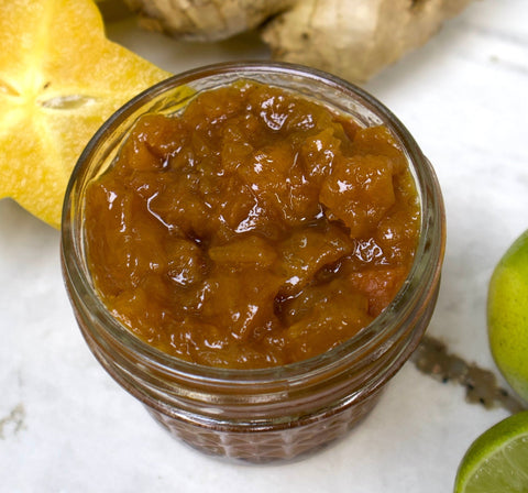 A small glass jar of iHeartFruitBox’s Star Fruit Ginger Preserve, chunky and amber-colored, is shown on a white surface with fresh starfruit, ginger, and lime nearby.