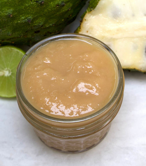 A glass jar of iHeartFruitBox Soursop Preserve, a light brown tropical spread, sits on a white surface with a halved soursop and lime in the background, emphasizing its fresh and vibrant appeal.