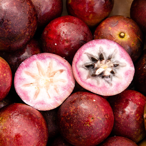 Close-up of multiple iHeartFruitBox Star Apples***Pre order*** with one split in half, revealing the white and black seeds inside. The outer skin is mostly dark purple with some lighter spots, making these tropical delicacies a visual and flavorful treat.